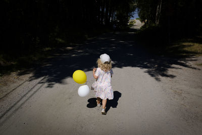 Close-up of cute girl running with balloons on road amidst trees