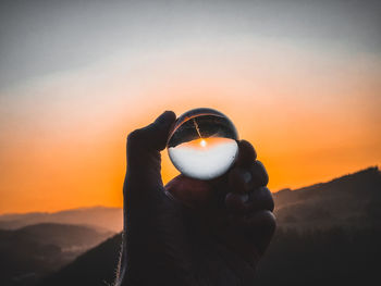 Close-up of hand holding orange against sky during sunset