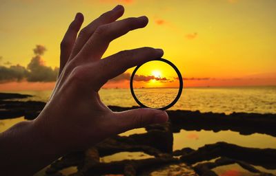 Close-up of human hand holding ring against sea during sunset