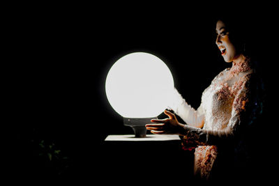 Side view of woman looking at illuminated lighting equipment against black background