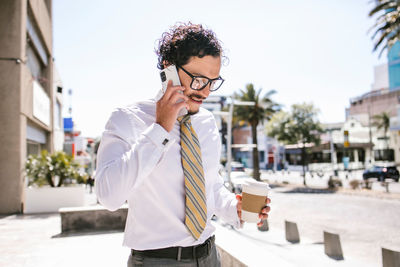 Positive male entrepreneur in formal suit and glasses looking away while having phone call on street while carrying a coffee in hand