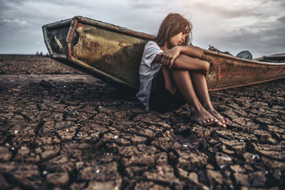 Sad girl sitting by boat on cracked land