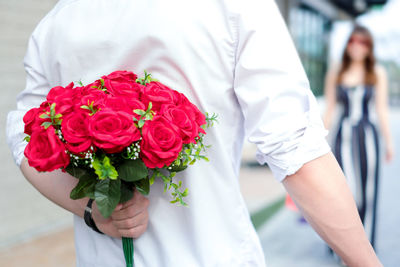 Midsection of man holding red roses with girlfriend in background