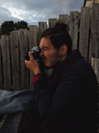 Side view portrait of young man holding camera while sitting outdoors