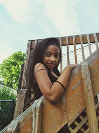 Portrait of smiling young woman sitting on railing against sky