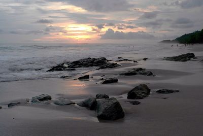 Rocks on wet beach against cloudy sky during sunset