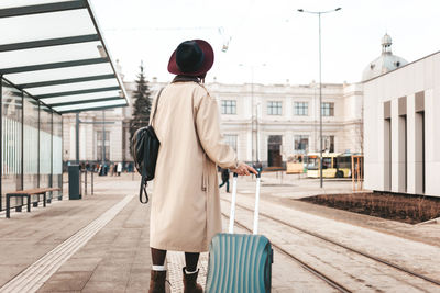 Stylish girl with a suitcase stands at a city stop awaiting a tram