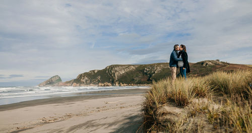 Side view of couple kissing while standing on beach against cloudy sky