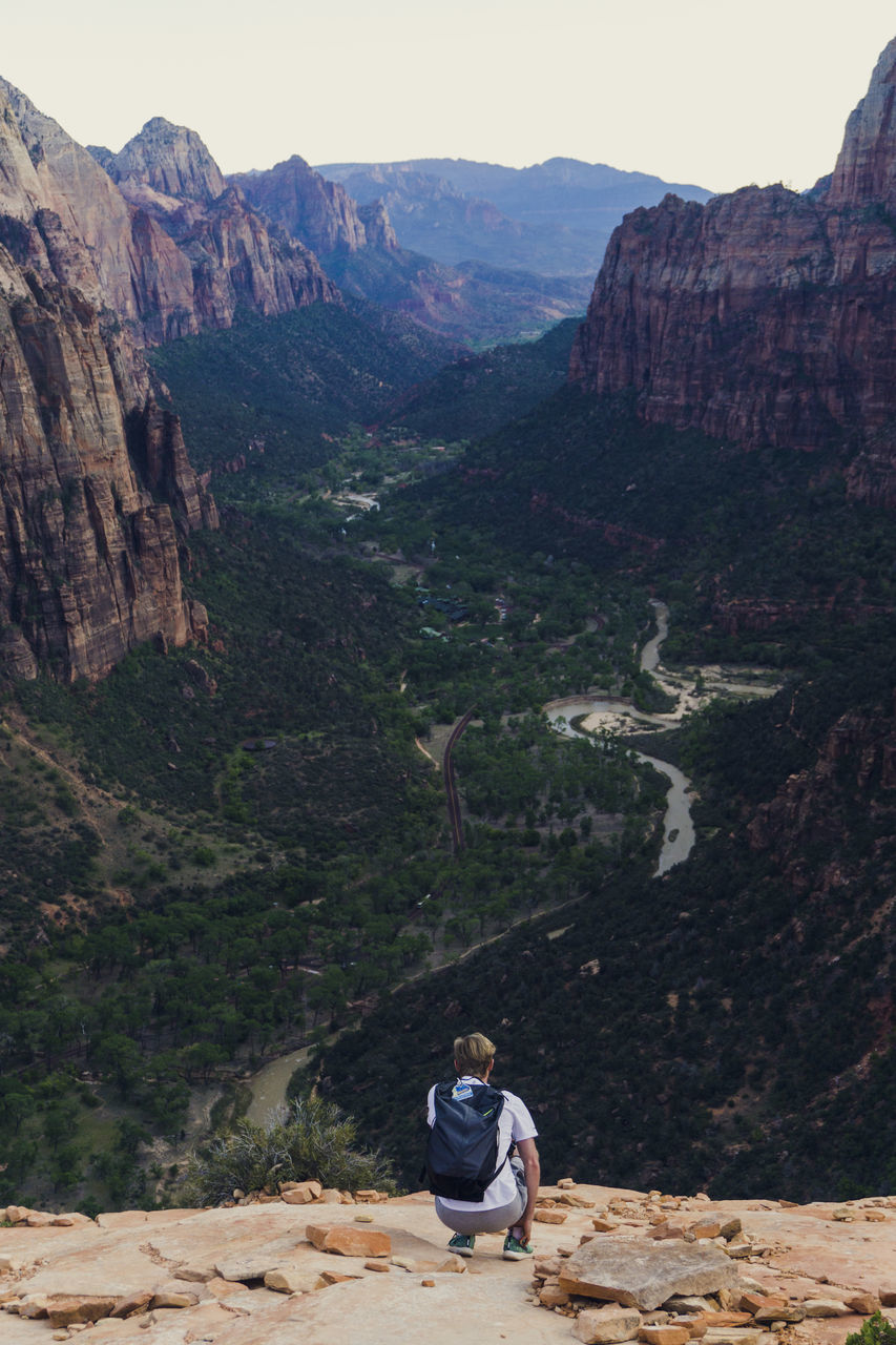 REAR VIEW OF MAN SITTING ON ROCK AGAINST MOUNTAIN