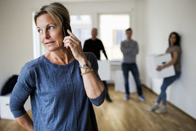 Mature woman using mobile phone while family carrying moving boxes in background at home