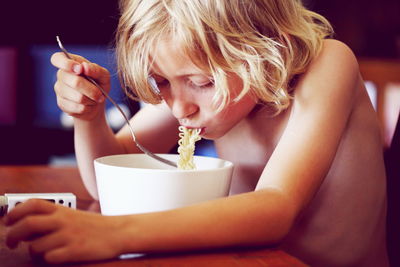 Shirtless blond boy eating noodles on table at home