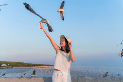 Smiling woman with hand raised feeding seagull while standing against sea