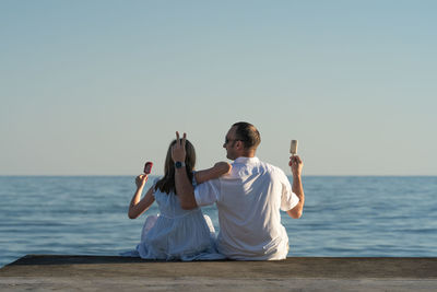 A girl and her dad are sitting on the pier and eating ice cream on a summer evening