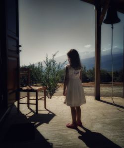 Rear view full length of girl standing by chair against sky