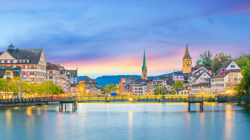 Illuminated buildings by river against sky in city