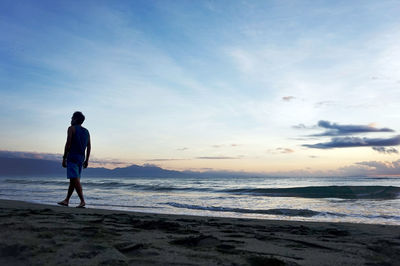 Rear view of man walking on shore at beach against sky during sunset