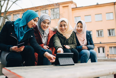 Happy female muslim friends sitting with digital tablet on bench in city