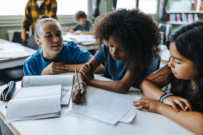 Professor crouching near female student writing on paper while sitting beside classmate in classroom at elementary schoo