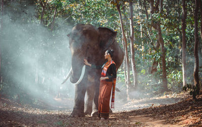 Full length of smiling woman standing with elephant in forest