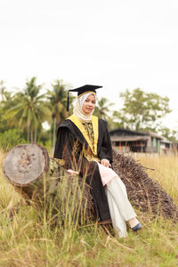 Young woman in graduation gown sitting on tree stump at field