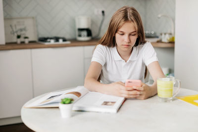 A teenage girl is sitting at a table in the kitchen, holding a phone and doing homework
