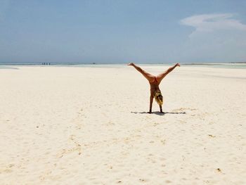 Rear view of young bikini woman doing handstand on sandy beach against sky