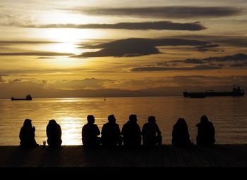 Silhouette people sitting by sea against sky during sunset