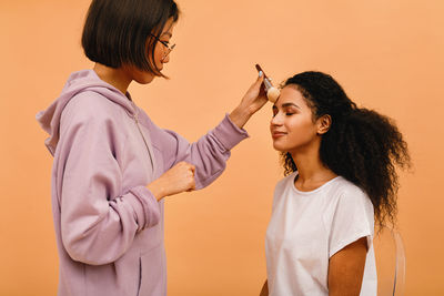 Woman doing make-up of woman against orange background