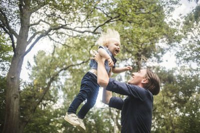 Low angle view of smiling man catching blond baby boy against trees