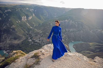 Brunette woman in a blue long dress stands on the edge of the sulak canyon in the evening at sunset