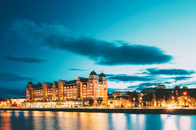 Illuminated buildings by river against cloudy sky