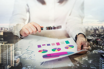 Double exposure of architect analyzing graphs using magnifying glass with city in background