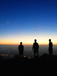 Silhouette friends standing on mountain by illuminated city against blue sky during sunset