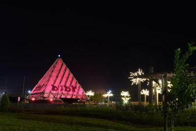 Illuminated house amidst trees and buildings against sky at night