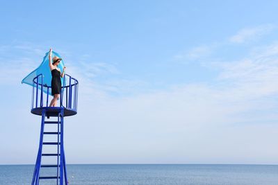 Young woman holding scarf on lookout tower by sea against sky