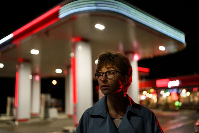 Depressed young woman in glasses stands alone near gas station looks away with sadness at cold night