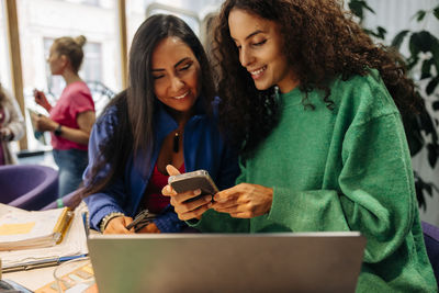 Smiling female colleague sharing smart phone with businesswoman at office