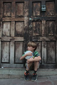 Full length of boy holding ball while sitting against closed doors