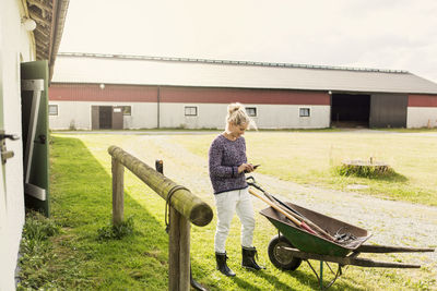 Woman using smart phone while standing by wheelbarrow at farm