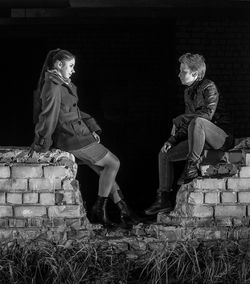 Full length of friends sitting broken brick wall at night