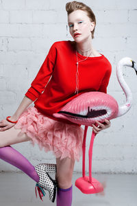 Beautiful woman holding red flamingo sculpture against wall