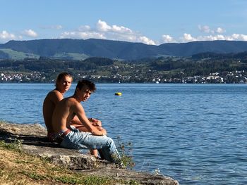 Shirtless man and boy sitting on rock by lake against sky