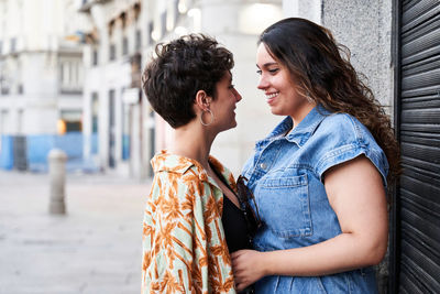 Glad young girlfriends in casual clothes smiling and looking at each other while embracing near building wall on street of madrid, spain