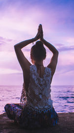Rear view of woman with arms raised meditating at beach against sky during sunset