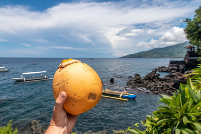 Cropped hand holding coconut by against cloudy sky