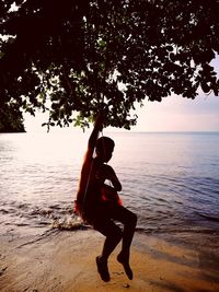 Silhouette of shirtless boy sitting on tree swing at beach during sunset