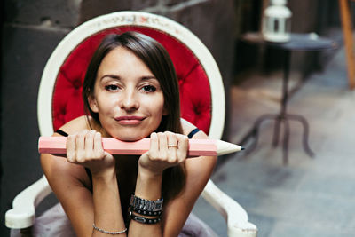 Portrait of beautiful woman holding large pencil while sitting on chair at home