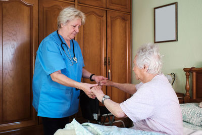 Side view nurse in uniform helping elderly female in nightwear to get out of bed with blanket in light bedroom at home