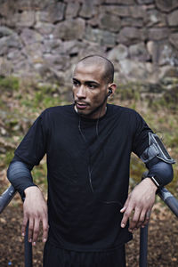 Thoughtful male athlete listening music while leaning on parallel bars in forest