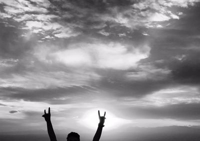 Cropped image of man showing rock sign against cloudy sky